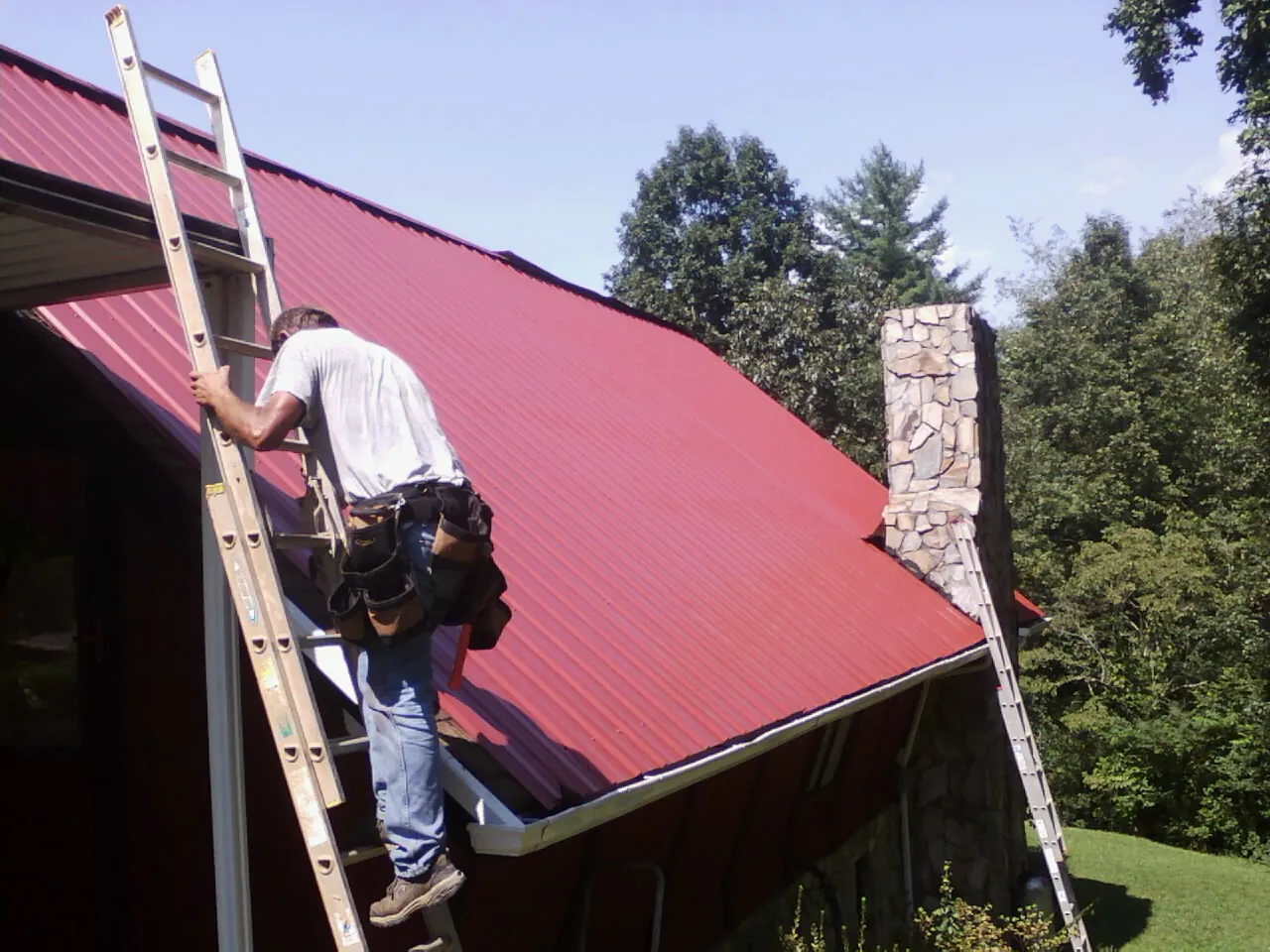 Roofer on a ladder beside a red metal roof and stone chimney — Soffit & Fascia Repair by Roofing Brothers in Dillwyn